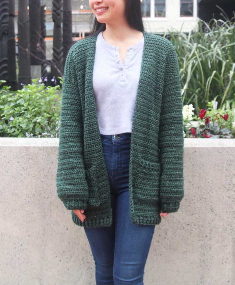 May include: A dark green crocheted cardigan with long sleeves and two pockets. The cardigan is open in the front and has a relaxed fit.