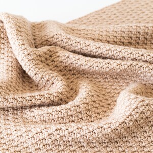 May include: A close-up of a beige crocheted blanket with a textured pattern. The blanket is folded and draped over a white surface.