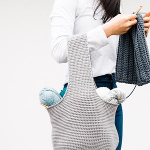 May include: A gray crocheted tote bag with a wide opening and two straps. The bag is filled with yarn and knitting needles.