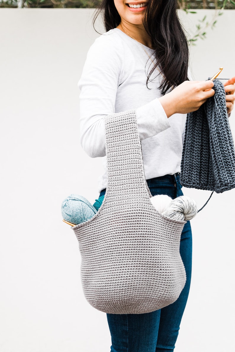 May include: A gray crocheted tote bag with a wide opening and two straps. The bag is filled with yarn and knitting needles.
