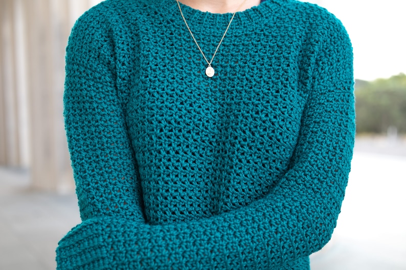 May include: A teal blue crocheted sweater with a delicate stitch pattern. The sweater has long sleeves and a crew neckline.