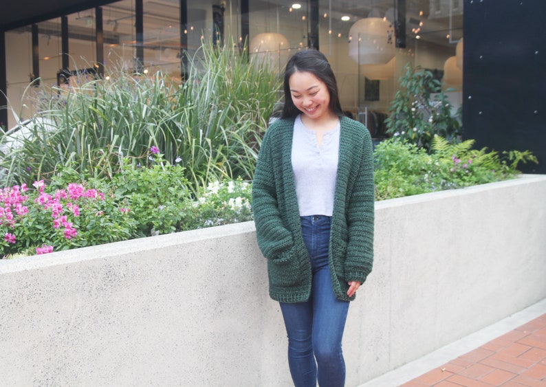 May include: A woman wearing a dark green crocheted cardigan with a white shirt and blue jeans. She is standing in front of a wall with plants.