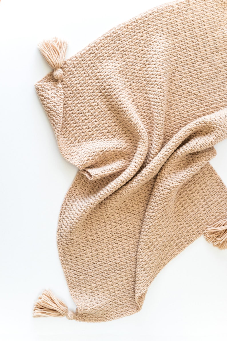 May include: A beige crocheted throw blanket with tassels on the ends. The blanket is folded and draped on a white surface.