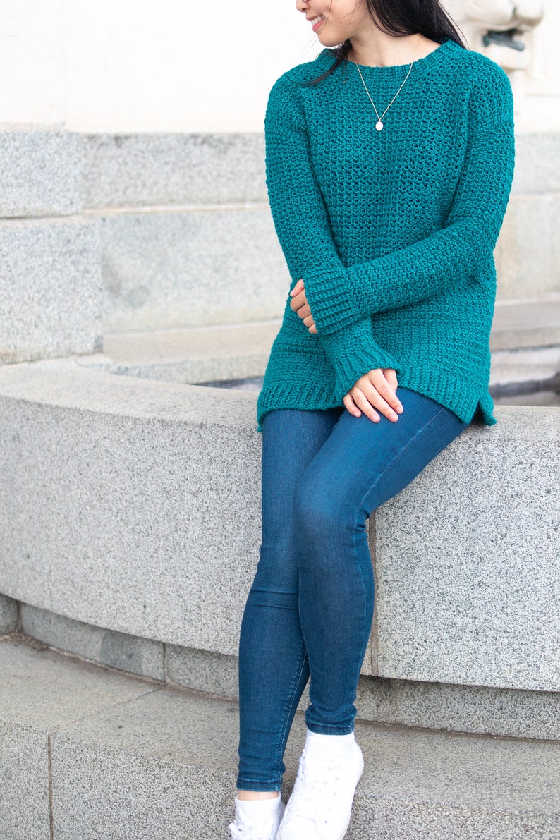 May include: A teal blue crocheted sweater with long sleeves and a relaxed fit. The sweater is worn by a person with blue jeans and white sneakers.