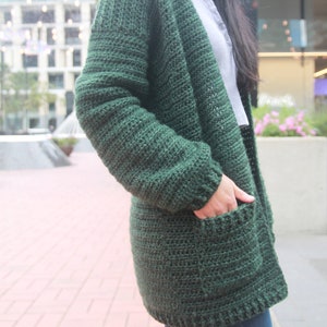 May include: A dark green crocheted cardigan with pockets. The cardigan is open in the front and has a long, loose fit.