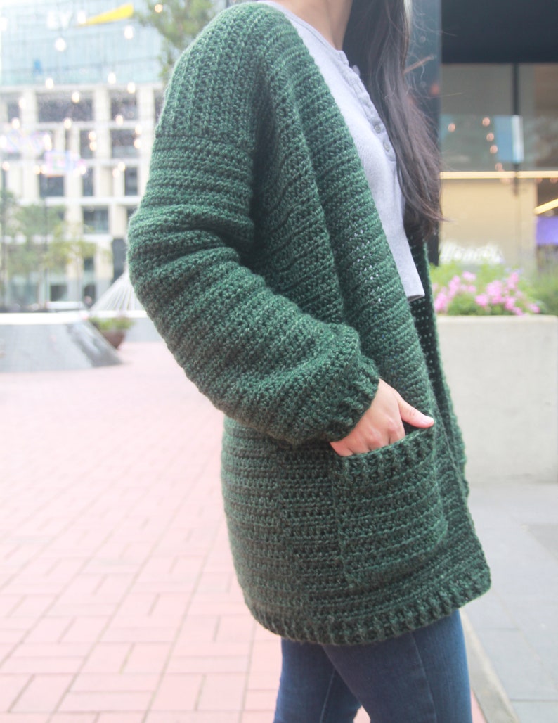 May include: A dark green crocheted cardigan with pockets. The cardigan is open in the front and has a long, loose fit.