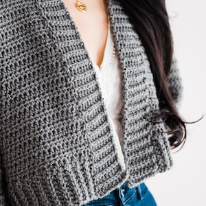 May include: A gray crocheted cardigan with a cropped hem, worn over a white top and blue jeans.