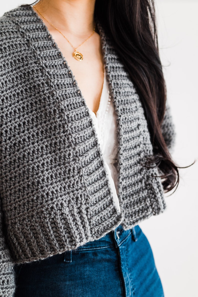 May include: A gray crocheted cardigan with a cropped hem, worn over a white top and blue jeans.