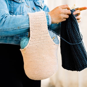 May include: A person wearing a denim jacket and black pants is holding a crocheted tote bag with yarn inside. The bag is beige and has a wide strap. The person is also holding a blue crocheted scarf.