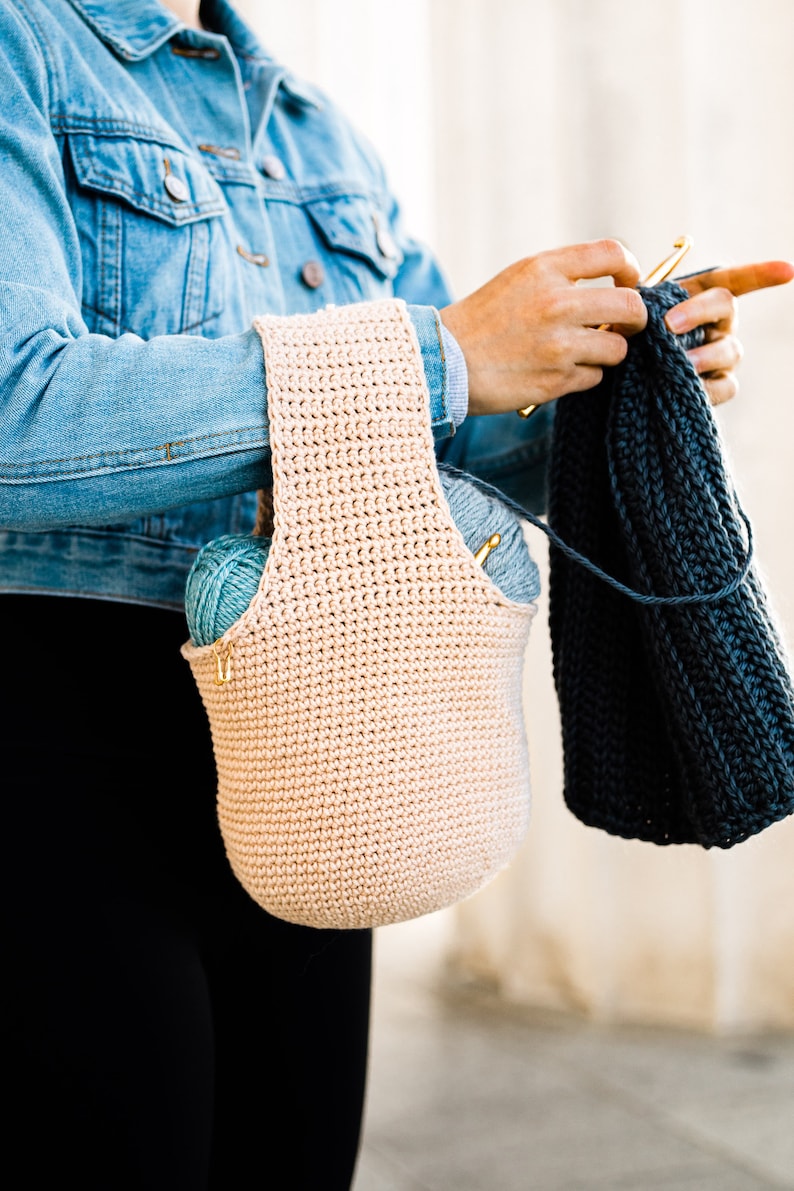 May include: A person wearing a denim jacket and black pants is holding a crocheted tote bag with yarn inside. The bag is beige and has a wide strap. The person is also holding a blue crocheted scarf.