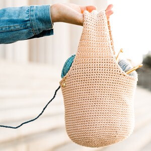 May include: A beige crocheted tote bag with yarn and knitting needles inside. The bag is being held by a hand wearing a denim shirt.