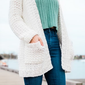 May include: A white crocheted cardigan with pockets and long sleeves. The cardigan is worn over a green crocheted top and blue jeans.