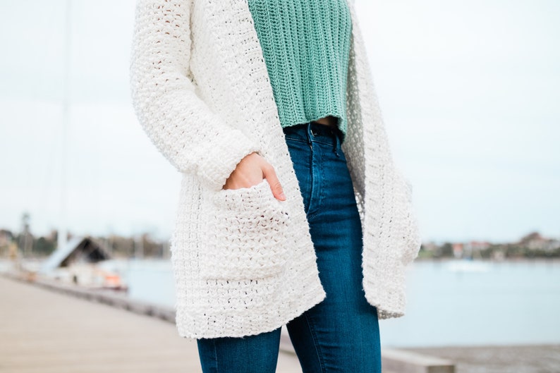 May include: A white crocheted cardigan with pockets and long sleeves. The cardigan is worn over a green crocheted top and blue jeans.