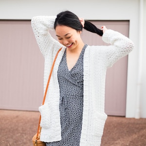 May include: A woman wearing a white crocheted cardigan with a blue floral dress and a brown woven bag.
