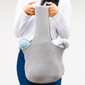 May include: A gray crocheted tote bag with two skeins of yarn inside. The bag has a wide, flat bottom and two long straps.