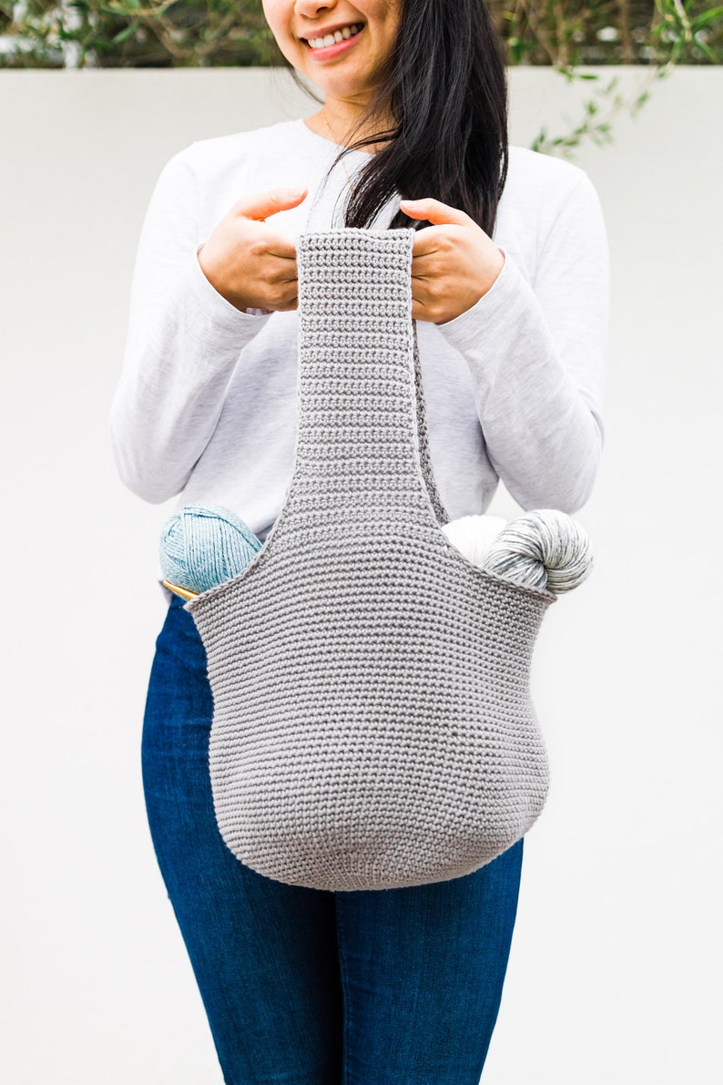 May include: A gray crocheted tote bag with two skeins of yarn inside. The bag has a wide, flat bottom and two long straps.