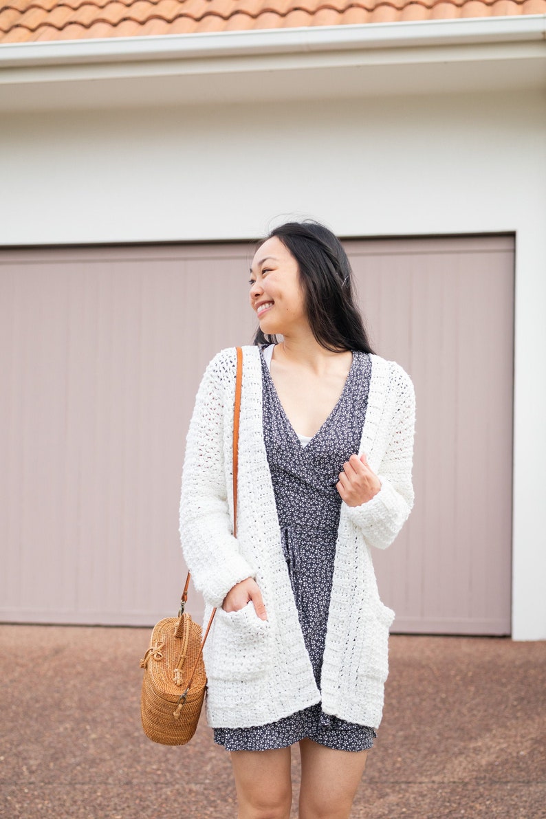 May include: A woman wearing a white crochet cardigan with a brown leather strap bag over her shoulder. She is wearing a floral print dress and smiling.