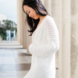 May include: A white crocheted cardigan with a loose fit and long sleeves. The cardigan is worn over a light pink tank top and blue jeans.