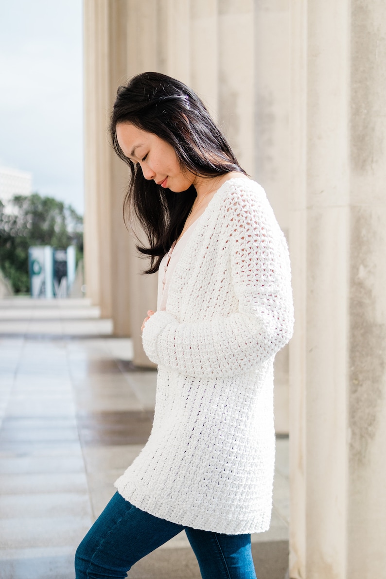 May include: A white crocheted cardigan with a loose fit and long sleeves. The cardigan is worn over a light pink tank top and blue jeans.