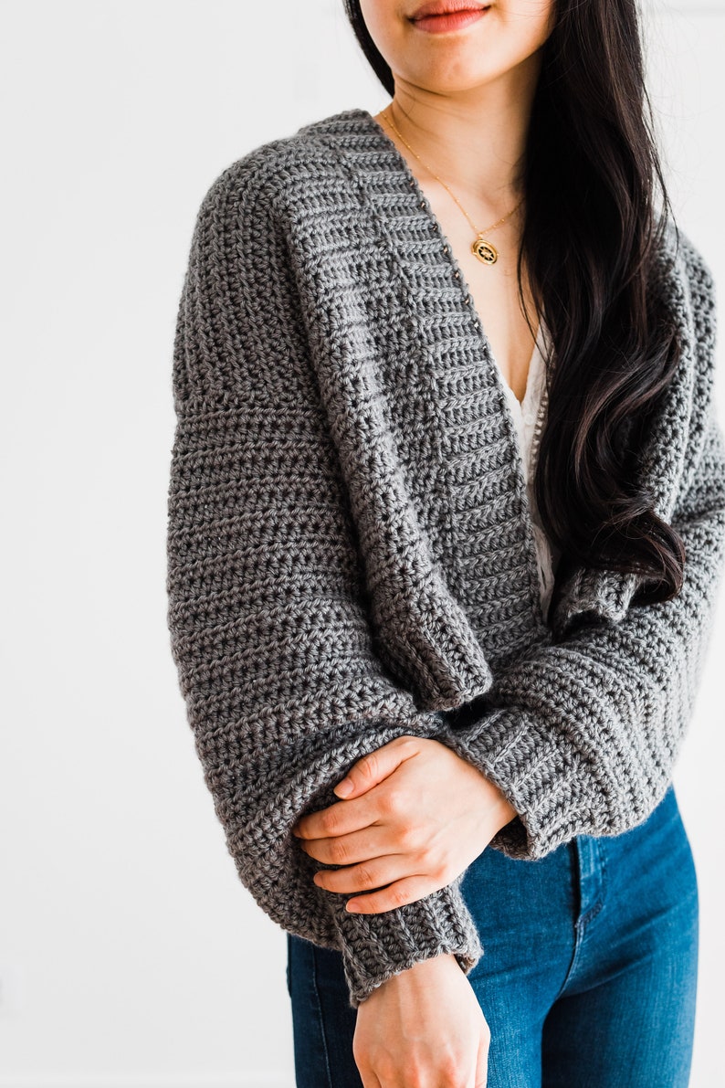 May include: A gray crocheted cardigan with long sleeves and a V-neck. The cardigan is worn by a person with long black hair.