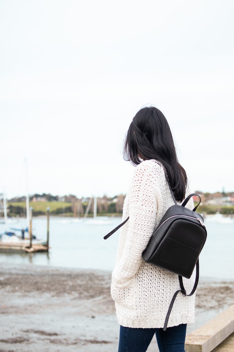 May include: A woman wearing a white crocheted cardigan and blue jeans is walking by the water. She is carrying a black leather backpack.