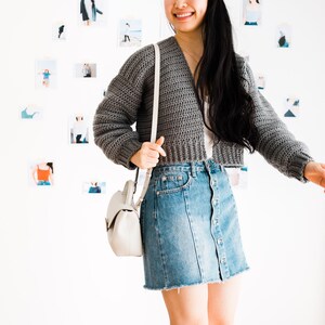 May include: A gray cropped cardigan with long sleeves and a V-neckline. The cardigan is worn over a white shirt and a denim skirt. The person is wearing a white crossbody bag.