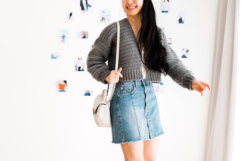 May include: A gray cropped cardigan with long sleeves and a V-neckline. The cardigan is worn over a white shirt and a denim skirt. The person is wearing a white crossbody bag.