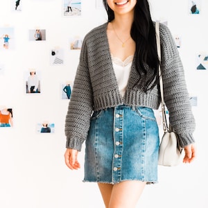 May include: A gray cropped cardigan with long sleeves and a V-neckline. The cardigan is worn over a white camisole and a denim skirt with button closures.