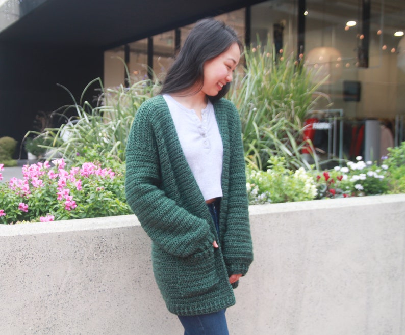 May include: A woman wearing a dark green crocheted cardigan with pockets. The cardigan is open in the front and the woman is wearing a white t-shirt underneath.