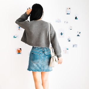 May include: A woman wearing a gray cropped sweater, a denim skirt, and white sneakers. She is standing in front of a white wall with several photos pinned to it.