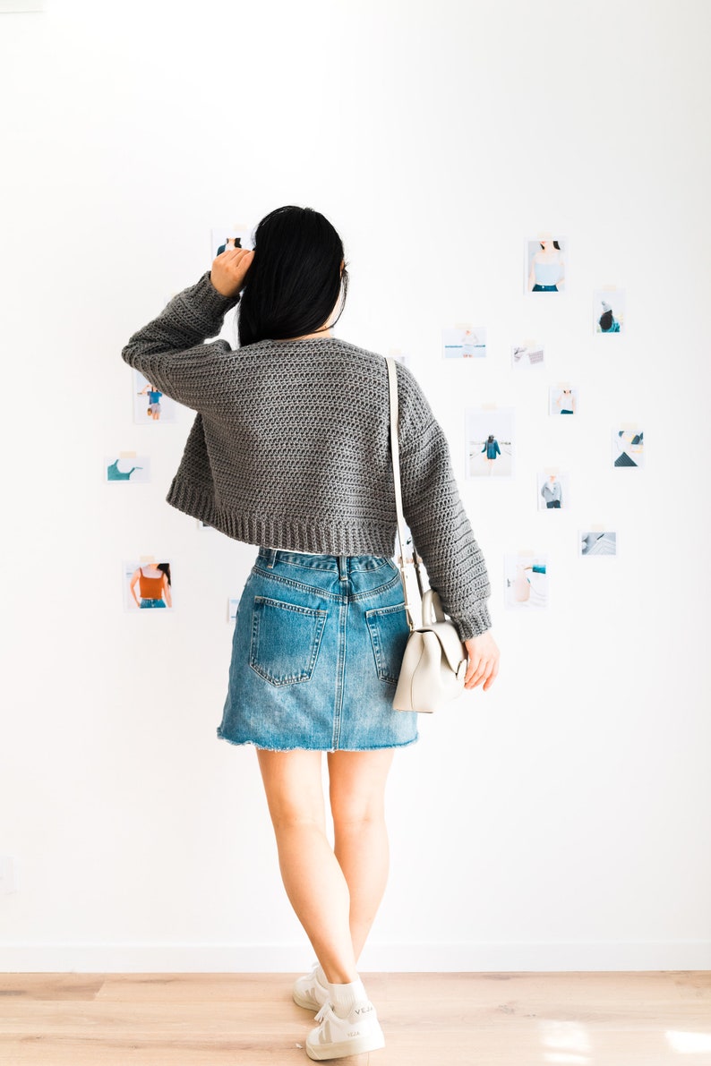 May include: A woman wearing a gray cropped sweater, a denim skirt, and white sneakers. She is standing in front of a white wall with several photos pinned to it.