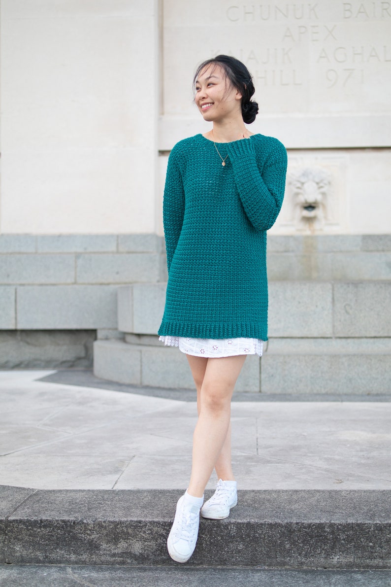 May include: A woman wearing a teal blue knitted sweater with a white skirt and white sneakers. She is standing on a set of stairs in front of a white building.