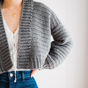 May include: A gray crocheted cardigan with a wide, open neckline and long sleeves. The cardigan is cropped and has a relaxed fit.