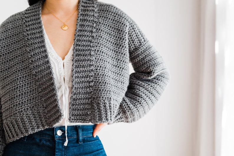 May include: A gray crocheted cardigan with a wide, open neckline and long sleeves. The cardigan is cropped and has a relaxed fit.