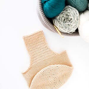 May include: A beige crocheted tote bag with a round bottom. The bag is lying on a white surface. There is a basket of yarn in the background.