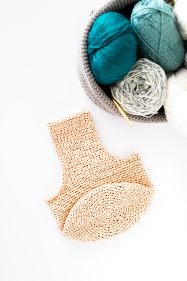 May include: A beige crocheted tote bag with a round bottom. The bag is lying on a white surface. There is a basket of yarn in the background.