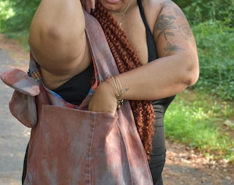 May include: A brown and blue tie-dye tote bag with a black strap. The bag is being held by a person with long, red braids.