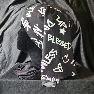 Könnte beinhalten: Ein schwarzes Kopftuch mit einem weißen und rosafarbenen Druck von Wörtern und Symbolen, darunter "Blessed", "Smile" und "Up".