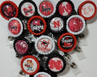 Peut inclure: Plusieurs enrouleurs de badges avec des motifs variés. Les enrouleurs présentent la lettre "N" et le mot "Huskers" en rouge et blanc, certains avec du texte supplémentaire comme "Go Big Red" et "Forever Huskers". Les enrouleurs ont des dessus en forme de bouchon de bouteille noirs, rouges et blancs.