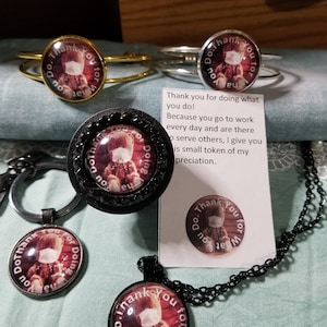 Könnte beinhalten: Sortiment an Schmuck mit einem runden Anhänger mit einem Teddybär, der eine Gesichtsmaske trägt. Der Anhänger lautet "Do Thank You for Doing What You Do".