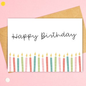 May include: A white birthday card with the words "Happy Birthday" in black script. The bottom of the card features a row of colorful candles with yellow flames. The card is inside a brown envelope, set against a pink background.