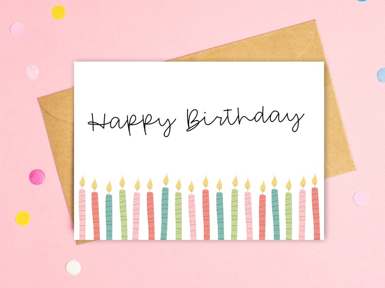 May include: A white birthday card with the words "Happy Birthday" in black script. The bottom of the card features a row of colorful candles with yellow flames. The card is inside a brown envelope, set against a pink background.