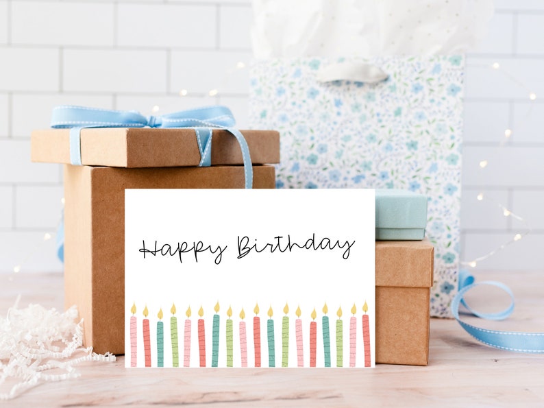 May include: A birthday card with the words "Happy Birthday" in black script, above a row of colorful candles. The card is in front of wrapped gifts and a gift bag, all in a light, airy setting.