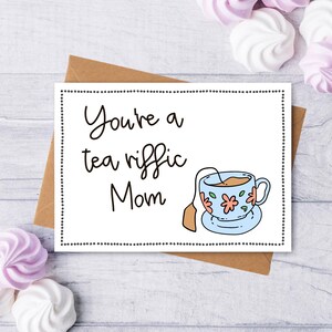 May include: A white card with black text that reads "You're a tea riffic Mom" and a cartoon illustration of a blue teacup with pink flowers and a tea bag.