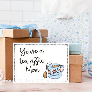 May include: A white card with black text that reads "You're a tea riffic Mom" and a cartoon illustration of a teacup with a tea bag. The card is sitting on top of a brown gift box.
