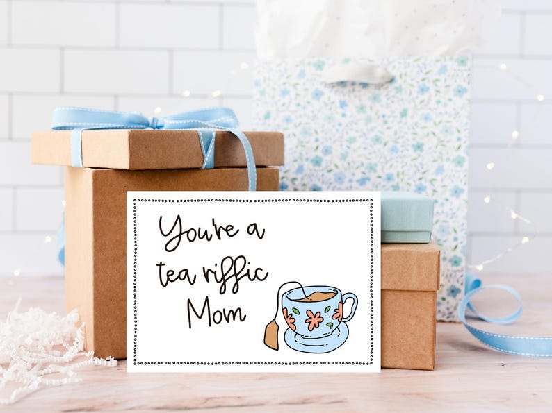 May include: A white card with black text that reads "You're a tea riffic Mom" and a cartoon illustration of a teacup with a tea bag. The card is sitting on top of a brown gift box.