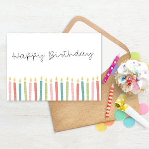 May include: A white birthday card with the words "Happy Birthday" in black script, featuring colorful candles at the bottom. The card is inside a brown envelope, with a cupcake, candles, and confetti.