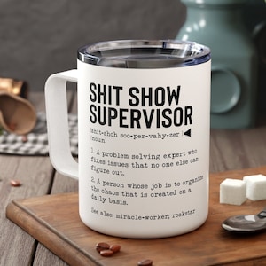 May include: White travel mug with a black and white design that reads "Shit Show Supervisor" with a definition of the term.