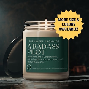 Puede incluir: Una vela blanca con una etiqueta verde que dice "The Sweet Aroma of A Badass Pilot". La etiqueta tiene un pequeño logotipo en la esquina inferior izquierda. La vela está sobre una superficie oscura.