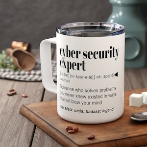 May include: White travel mug with a black design that reads "cyber security expert" and a definition of the term. The mug is sitting on a wooden surface with coffee beans scattered around it.